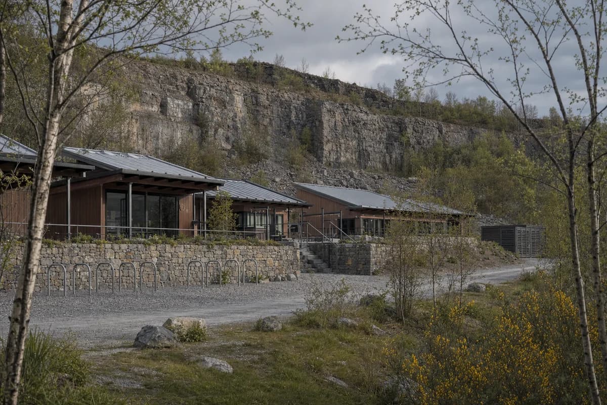 Unoccupied low stone and chestnut climbing-center pavilions at a regenerating quarry edge.