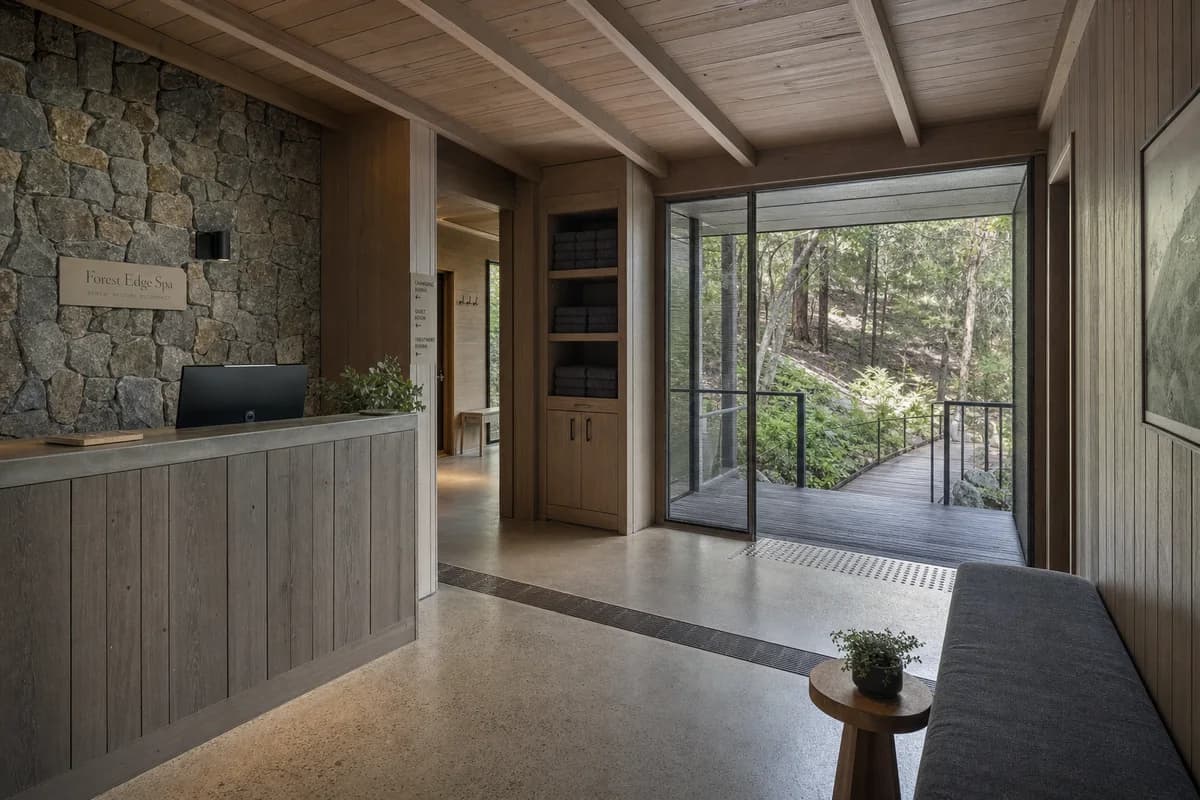 Unoccupied weathered-timber spa reception opening through a screened threshold to managed woodland.