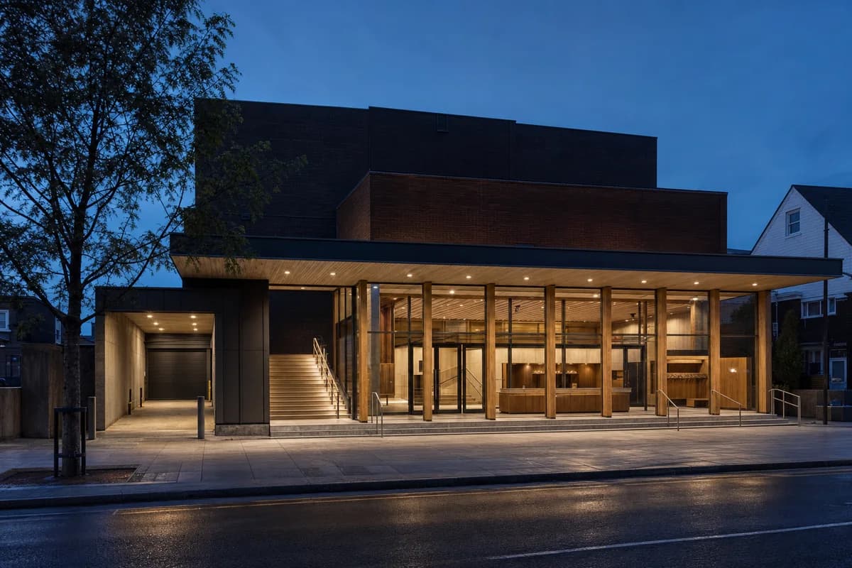 Dark brick puppet theater with a timber-lit foyer at blue hour.