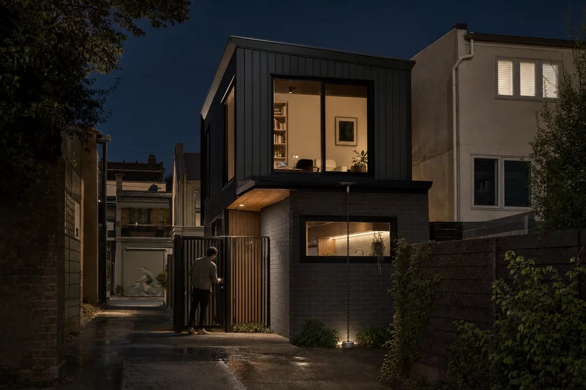 Compact dark masonry laneway house with restrained entry and interior lighting.
