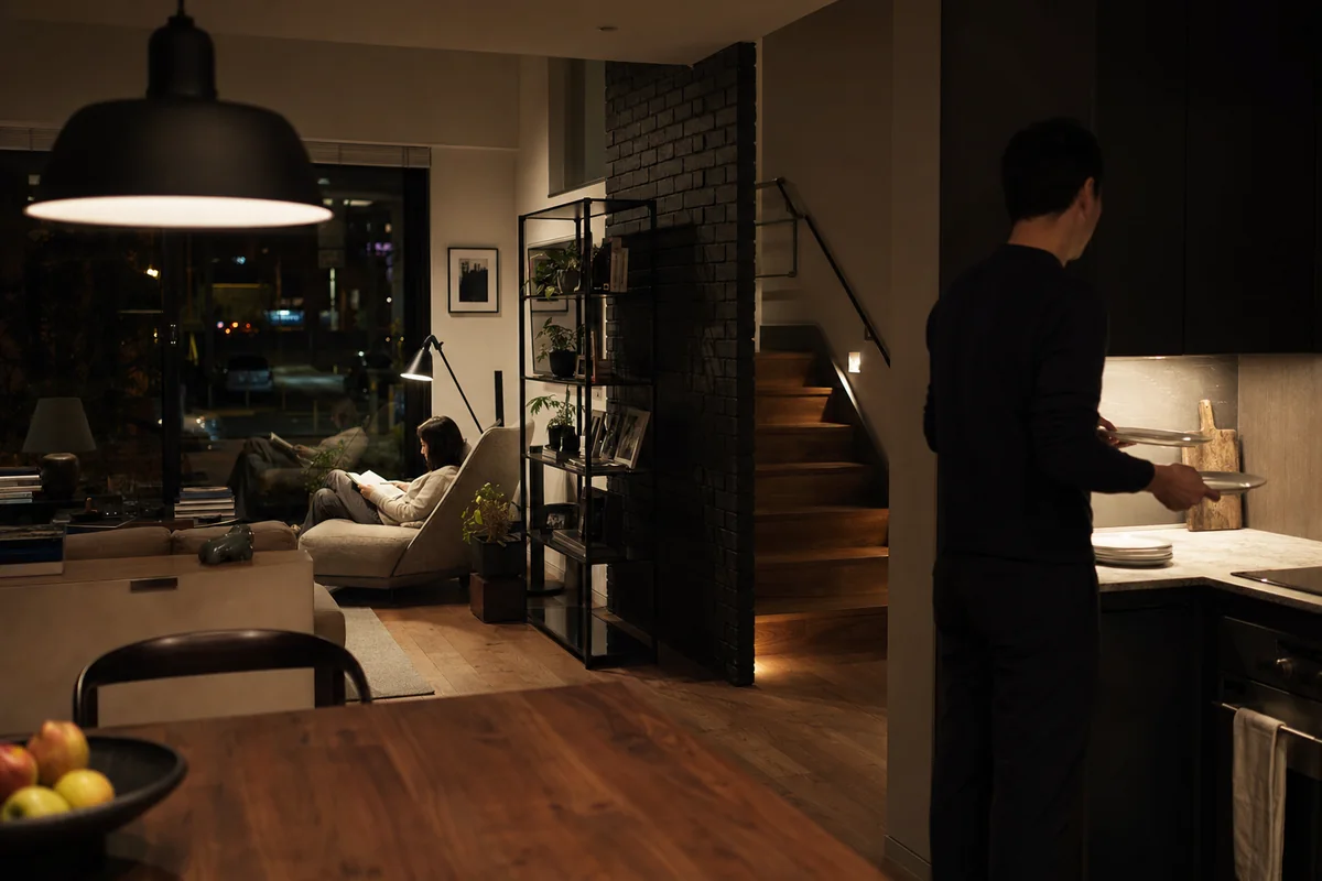 Dark townhouse living room with restrained lamps and two residents at evening routines.